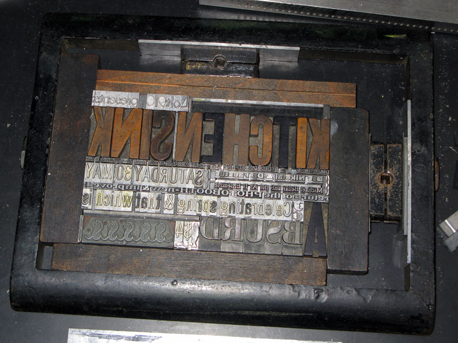 An image of letterpress type locked up and waiting for printing. Large wood letters read (backwards) KITCHENSINK. 