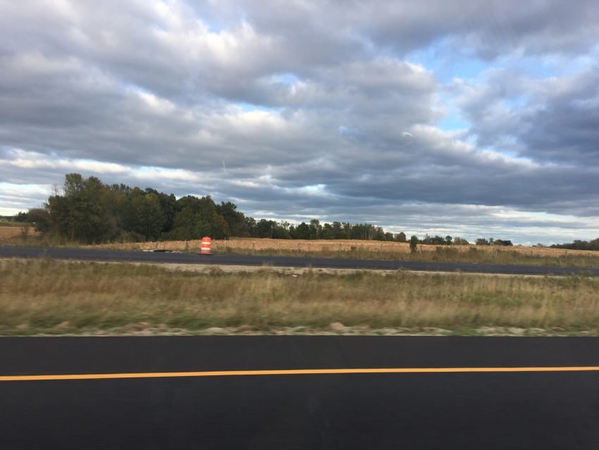 An image of the landscape from the window of a car going down the interstate. There are clouds, distant trees, and a small orange traffic construction cone in the background.
