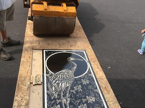 A carved and inked wood block with an image of a heron on it sits on the ground waiting for paper. Behind it a streetroller waits to press it.