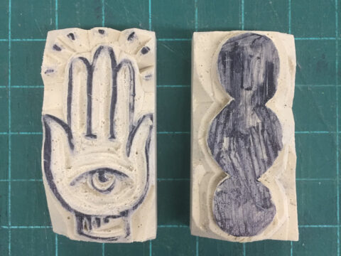An image of two small erasers carved for relief printing. One eraser shows the hamsa, a symbol with an eye in a hand. The other eraser shows three circles touching.