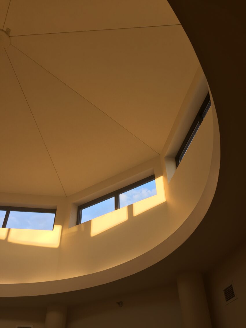 An image of the ceiling of the second floor of the Bush Art Center. There are rectangular windows set into a circular large cupola-type space. It is late in the day and the yellow light comes through at a slant.