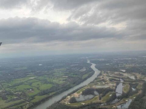 An aerial image of a river cutting through land outside of Nashville, TN. The clouds are gray and backlit. The land in the distance is hazy and blue. The land up close is brown and green. It is spring.