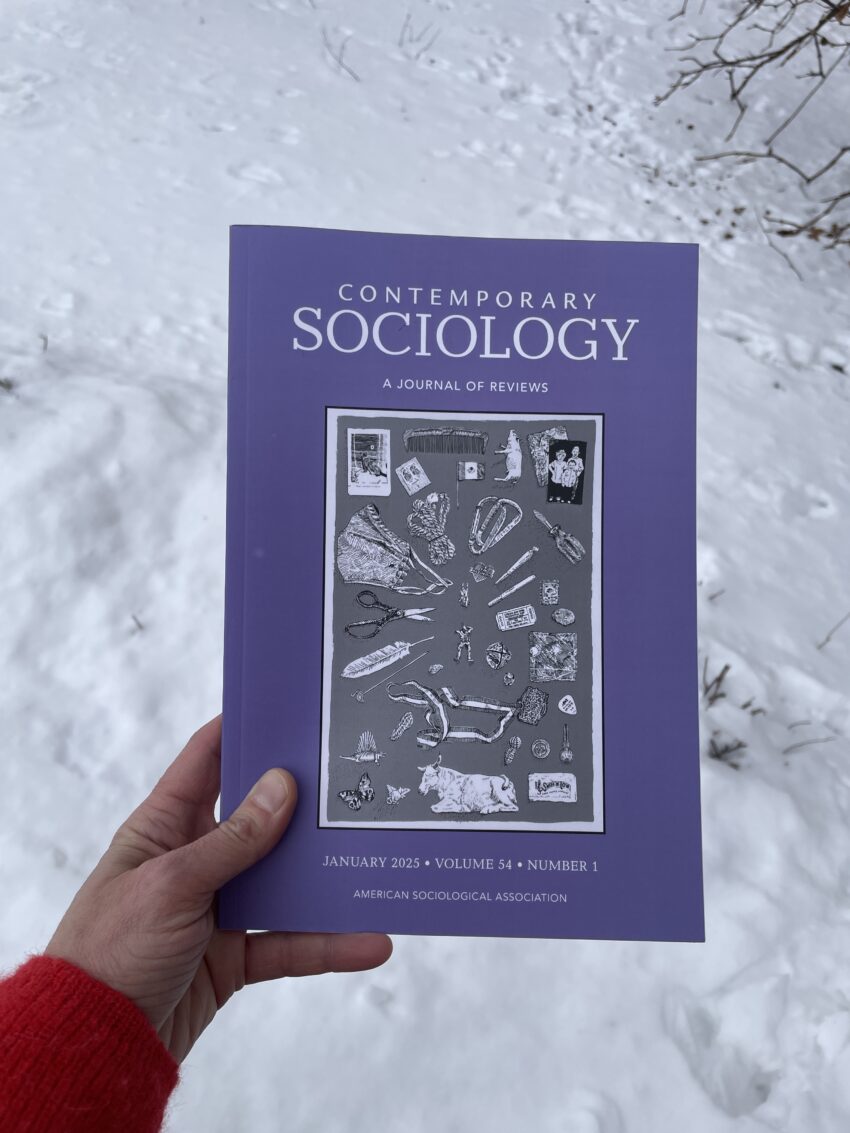 An image of a white hand holding a print copy of the journal "Contemporary Sociology." The cover of the journal features a black and white image of a print by Katie Ries. The print is a line drawing of several small objects.